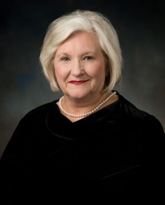 An older woman with short gray hair, wearing a black top and a pearl necklace, poses in front of a dark, neutral background.