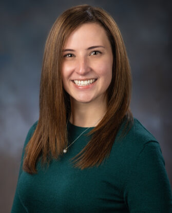 A woman with straight brown hair wearing a green long-sleeve top and a necklace, smiling at the camera against a blurred studio background.
