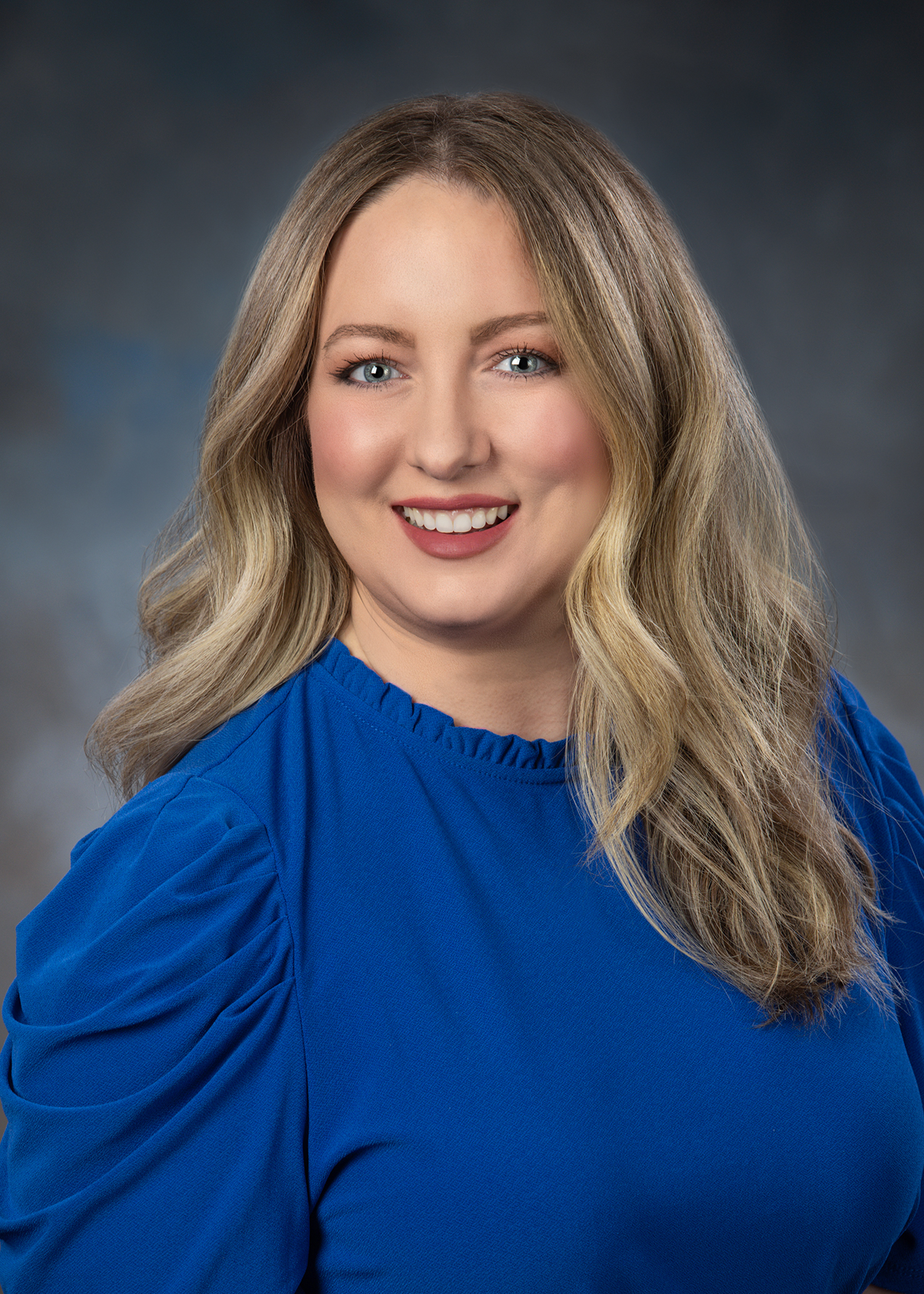 A woman with long, wavy blond hair wearing a bright blue blouse smiles at the camera against a neutral, softly blurred background.