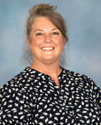 A smiling woman with grey-streaked hair pulled up, wearing a dark blue top with a white floral print.