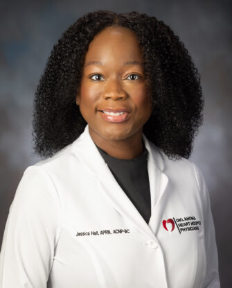 A woman with curly hair, wearing a white medical coat with the logo Oklahoma Heart Hospital Physicians and a black shirt, poses for a professional headshot against a neutral background.