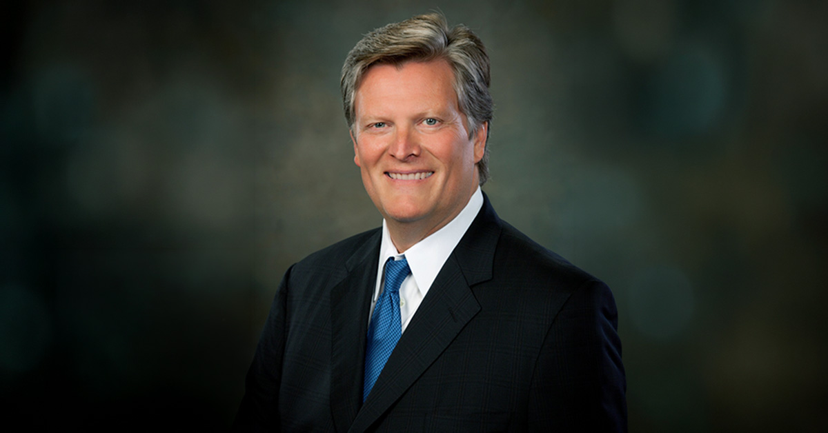 A man in a dark suit, blue tie, and light shirt is smiling at the camera against a dark, blurred background.