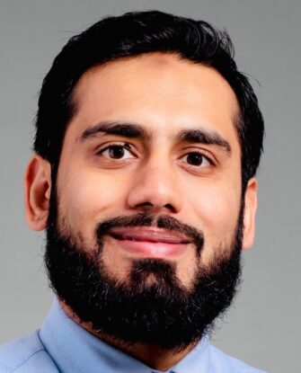 A man with dark hair and a beard, wearing a light blue collared shirt, smiles at the camera against a plain gray background.