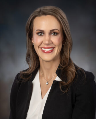 A woman with long brown hair wearing a black blazer over a white blouse, smiling at the camera against a dark, blurred background.